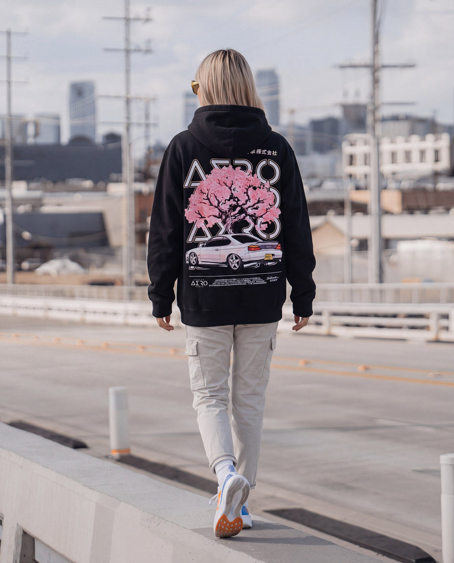 Woman walking across a bridge in a black hoodie with a pink cherry blossom and S15 print on the back