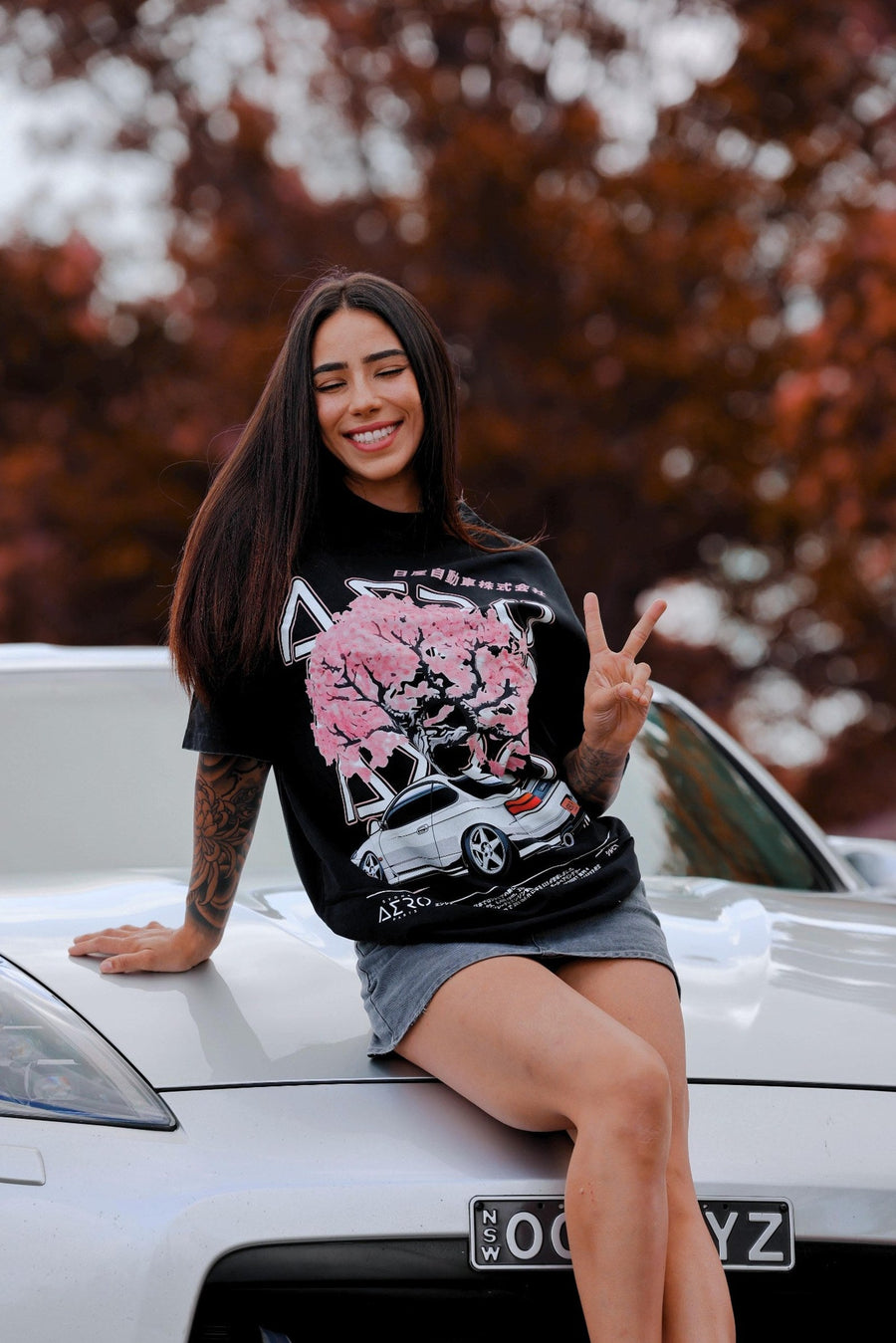 Woman sitting on a silver car bonnet in the Oversteer tee, a black oversized tee with a pink blossom tree and white S15 Silvia print