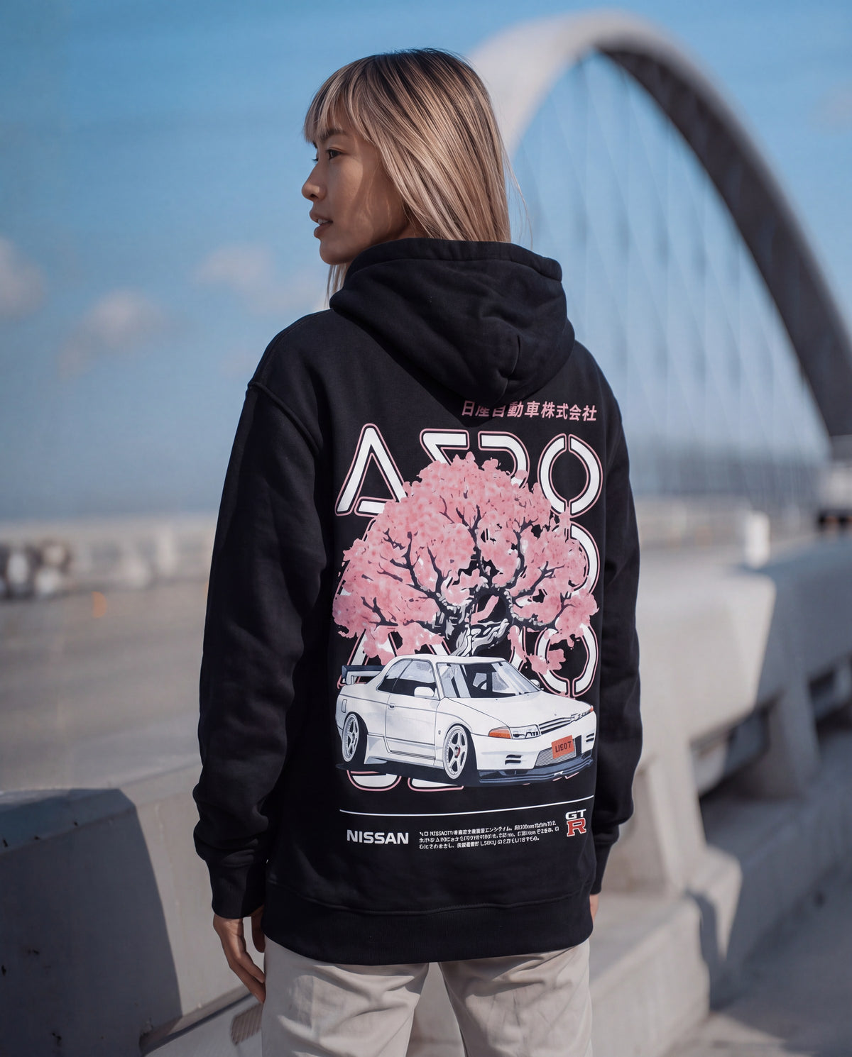 Woman on a bridge in a black hoodie with a pink blossom tree and white R32 GT-R back print