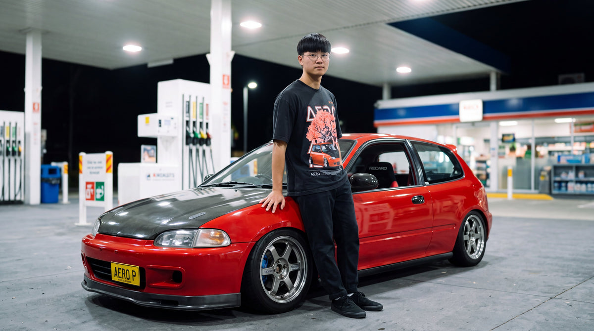 The Redlining Tee at a petrol station at night, with a red EG Civic behind him