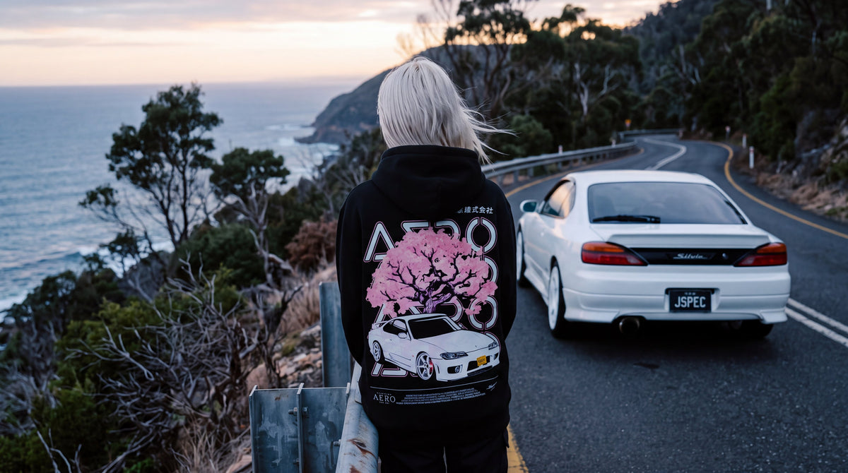 The Touge Hoodie worn at a coastal guardrail at dusk, with a white S15 Silvia on the road beside her