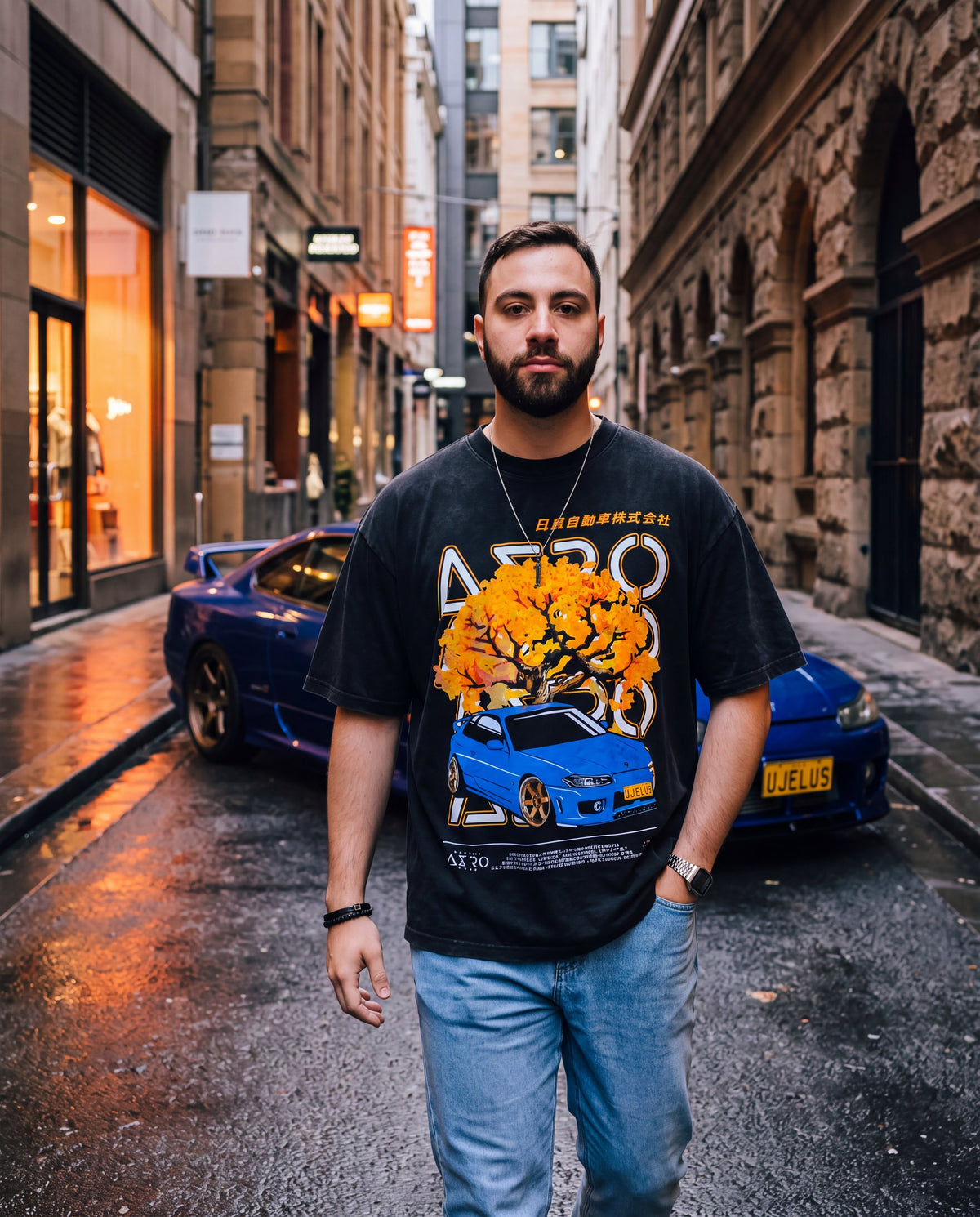 The UJELUS Tee in a wet Sydney sandstone laneway, orange blossom and blue S15 print on the front, with a blue S15 Silvia on bronze wheels behind him