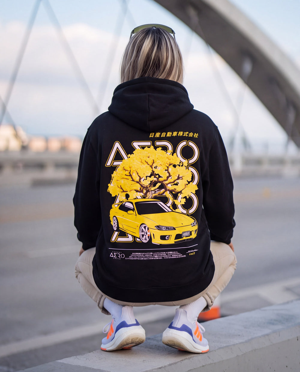 Woman crouched on a bridge barrier in a black hoodie with a yellow blossom tree and yellow S15 Silvia back print